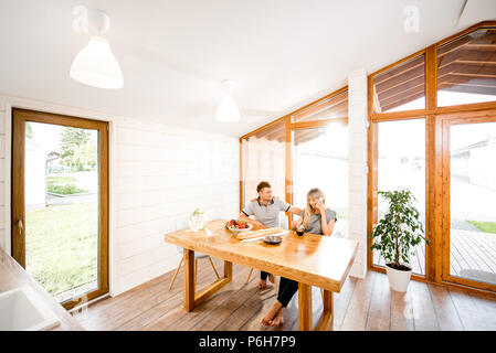 Junge und glückliche Paar mit einem Frühstück an der hölzernen Tisch sitzen in der modernen Landhaus mit großem Fenster in den Hintergrund Stockfoto