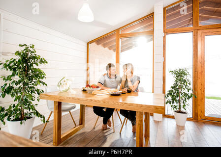 Junge und glückliche Paar mit einem Frühstück an der hölzernen Tisch sitzen in der modernen Landhaus mit großem Fenster in den Hintergrund Stockfoto
