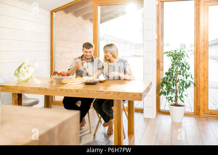 Junge und glückliche Paar mit einem Frühstück an der hölzernen Tisch sitzen in der modernen Landhaus mit großem Fenster in den Hintergrund Stockfoto