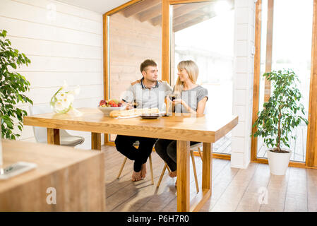 Junge und glückliche Paar mit einem Frühstück an der hölzernen Tisch sitzen in der modernen Landhaus mit großem Fenster in den Hintergrund Stockfoto