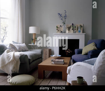 Weißen und blauen Sesseln und ein graues Sofa seta um einen niedrigen hölzernen Tisch vor dem Kamin in ein weißes Wohnzimmer Stockfoto