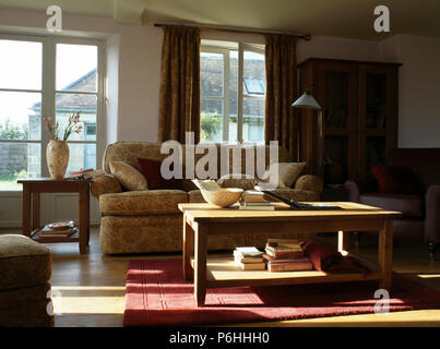 Bücher, die auf einfachen Holz Zwei-klassen-Tabelle im Land Wohnzimmer mit beige gemusterten Sofa vor Windows Stockfoto