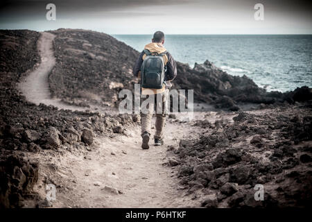 Einsamen Reisenden jungen Mann mittleren Alters mit Trekking Kleidung und Rucksack wandern auf dem Ozean Küste in einem Felsen Wüste. Nach einem Pfad mit vi Stockfoto