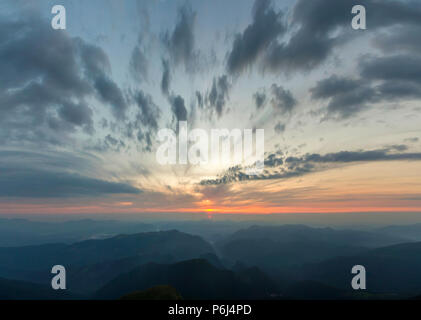 Fantastischer Sonnenaufgang oder Sonnenuntergang über grünen Bergrücken mit dichten blauen Nebel bedeckt. Helles orange sun Erhöhung in weichem bewölkter Himmel über Horizont. Stockfoto