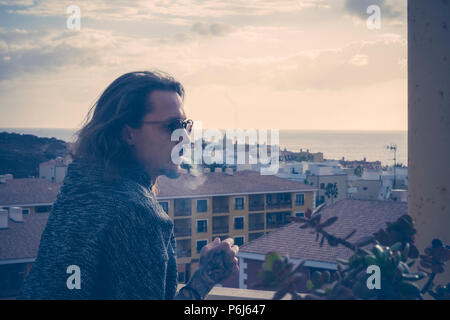 Ernsthafte schöner Mann Modell bleiben draußen auf der Terrasse zu Hause mit schönen Blick auf die Häuser und die Stadt und das Meer Horizont. Sonnenuntergang im Sommer Nacht. smok Stockfoto