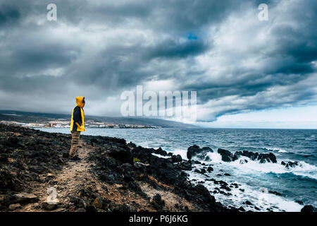 Mann mit gelben Jacke in einem wilden Platz auf dem Ozean Küste suchen die Kraft der Wellen in einem schlechtem Wetter. Herausforderung und genießen die Reisen Lifestyle Stockfoto