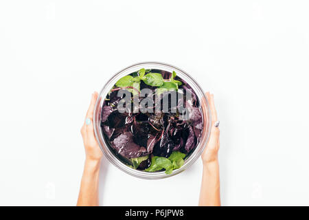 Woman's Hände halten große Glasschale mit Wasser gefüllt, lila Basilikum und Pfefferminze, Ansicht von oben. Minimale Anordnung der Gruenen waschen, flach. Stockfoto