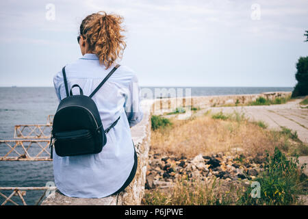 Ansicht der Rückseite des junge Frau mit Rucksack auf Attika mit Blick auf das Meer. Weibliche Reisende nachdenklich auf die Küste. Stockfoto