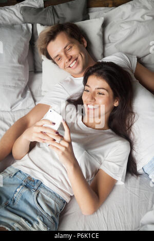 Lächelnden jungen Paar im Bett liegend das Lesen von Nachrichten auf dem Smartphone Stockfoto