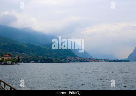 Comer See mit Gebäuden auf Küste und Alpen Berg mit Wolken im Hintergrund. Stockfoto