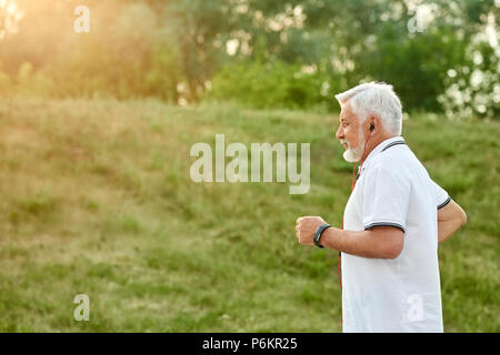 Alter Mann in Green City Park mit verschiedenen Pflanzen und Gras auf den Sonnenuntergang. Tragen klassisches weißes Poloshirt mit dunkelblauem Streifen, Sportuhr und rote Kopfhörer. Suche sportlich, gesund. Stockfoto