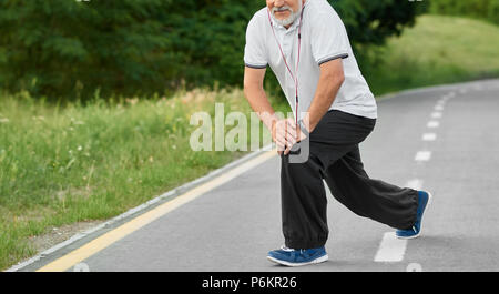 7/8 Foto der Ausbildung älterer Mann mit weißem Bart, white Polo Shirt, schwarze Hosen und blaue Turnschuhe. Dabei exrcises auf Rennstrecke im City Park. Gesunder Lebensstil , sportliche Figur. Stockfoto