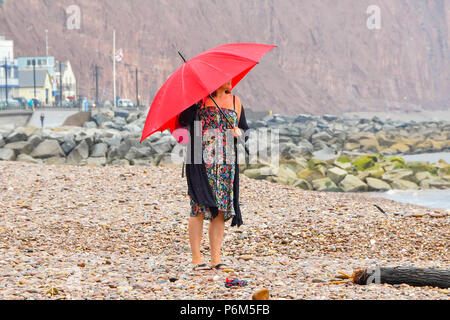 Honiton, Devon, Großbritannien. 1. Juli 2018. UK Wetter. Eine Frau auf dem Strand im Badeort Sidmouth Devon mit Schirm, der als die Stadt fängt den Rand eines vorbeifahrenden heftiger Regenschauer. Foto: Graham Jagd-/Alamy leben Nachrichten Stockfoto