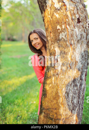 Junge Frau im rosa Kleid peekaboo, hinter dem Baum und lächelnd an sonnigen Frühlingstag. Stockfoto