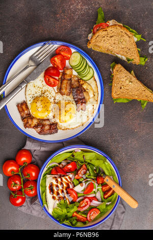 Essen flach: Salat mit Ziegenkäse, Sandwiches mit Käse und Gemüse und gebratene Eier mit Speck. Schönes helles Frühstück auf einem dunklen Stockfoto