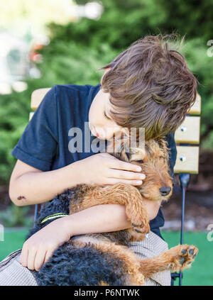 Junge sitzt auf einem Stuhl und Umarmen einen kleinen Welpen, Hund - Welsh Terrier zu schließen. Liebe portrait. Mit Haustiere und Tiere der Kindheit. Stockfoto