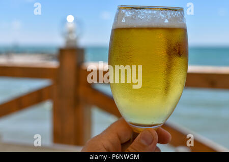 Toast mit einem Verschwitzten Tasse kaltes Bier an einem heissen Sommertag in einem Restaurant am Meer Stockfoto