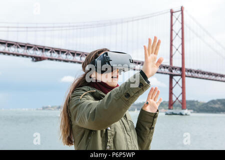 Junge schöne Mädchen mit Virtual reality Brillen. 25 April Brücke in Lissabon im Hintergrund. Das Konzept der modernen Technologien und deren Einsatz im täglichen Leben Stockfoto