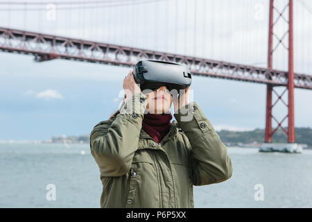 Junge schöne Mädchen mit Virtual reality Brillen. 25 April Brücke in Lissabon im Hintergrund. Das Konzept der modernen Technologien und deren Einsatz im täglichen Leben Stockfoto