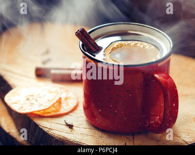 Glühwein in einem roten Keramik Becher über rustikalen hölzernen Brettern umgeben Gewürze. Stockfoto