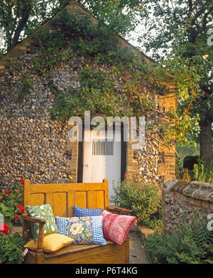 Bunte Kissen auf hölzernen Sitzbank im Garten vor der Feuerstein + Ziegel Cottage Stockfoto