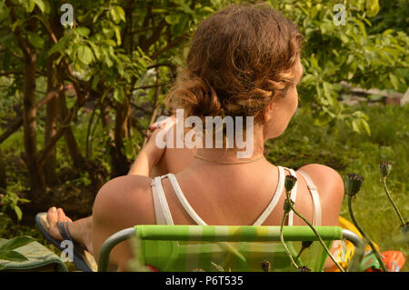 Eine junge Frau mit dem Lockigen blonden Haar in einem weißen T-Shirt in einem Stuhl im Garten ausruhen Stockfoto