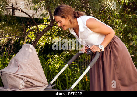 Eine junge dunkelhaarige Frau in einem weißen Hemd beige Rock ist zu Fuß mit einem kleinen Baby in einem Kinderwagen im Sommer heller Tag im Park Stockfoto