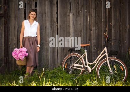 Eine junge dunkelhaarige Frau in einem weißen Hemd beige Rock ist mit einem schönen Blumenstrauß rosa Pfingstrosen in einem Weidenkorb. Weiter ist eine beige Retro b Stockfoto