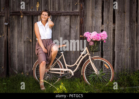 Eine junge dunkelhaarige Frau in einem weißen Hemd beige Rock ist mit einem schönen Blumenstrauß rosa Pfingstrosen in einem Weidenkorb. und sitzt auf einem beige ret Stockfoto