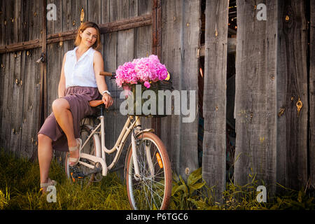 Eine junge dunkelhaarige Frau in einem weißen Hemd beige Rock ist mit einem schönen Blumenstrauß rosa Pfingstrosen in einem Weidenkorb. und sitzt auf einem beige ret Stockfoto