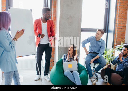 Junge Menschen verschiedener Rassen und Nationalitäten in Team building Training, mit Blick auf die afrikanischen Mann seine Ideen präsentieren auf Flipchart bei Sunny lof Stockfoto