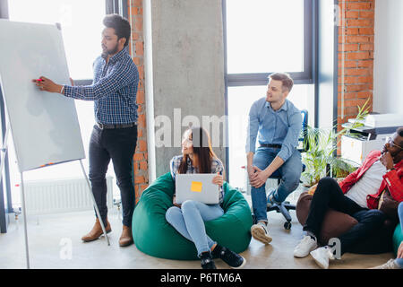 Hübscher junger indischer Geschäftsmann eine Präsentation auf einem Flipchart zu seinem aufmerksamen Mitarbeiter sitzen in gut beleuchteten Sitzungszimmer des Büro für Gestaltung Stockfoto