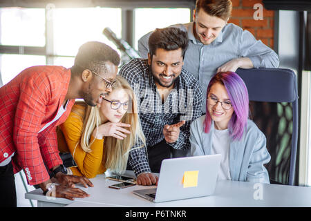 Multiethnischen fröhliche junge männliche und weibliche Studenten, die sich um die Tabelle in Campus ansehen Fotos mit einem Laptop für alle. Stockfoto