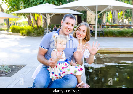 Porträts von Happy Family von drei - Vater, Mutter und Tochter schwenkten ihre Hände in die Kamera in der Nähe von Teich im Park sitzen. Familie die Zeit t Stockfoto