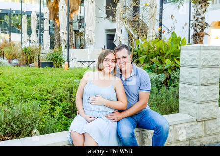 Portrait von Erwachsenen glücklich umarmte schwanger Familie, Paar beim Spaziergang im Stadtpark posieren. Fruchtbarkeit Konzept. Abendlicht. soft selektiven Fokus, Co Stockfoto