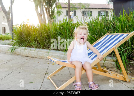 Portrait von niedlichen emotionale blondy Kleinkind Mädchen in weißem Kleid sitzt auf dem Liegestuhl und Schreien. City Park Recreation Area. Selektiver Fokus, Kopieren s Stockfoto