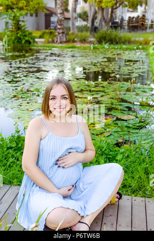 Glückliche junge Schöne schwangere Frau im blauen Kleid berühren Ihr Bauch mit Liebe und Sorgfalt sitzen in der Nähe von Teich mit Seerosen im Park. Weiche selecti Stockfoto