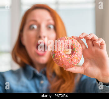 Rothaarige Frau mit Donut zu Hause sehr glücklich und aufgeregt, Sieger Ausdruck feiern Sieg schreien mit einem großen Lächeln und erhobenen Händen Stockfoto