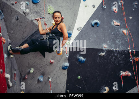 Kompromisslos sportliche Schüler Mädchen hängen mit Sicherungsseil und Kabelbaum Luft auf künstliche Felsen, Blick nach unten, auf der Suche nach einem Weg nach unten zu erhalten. Farben Rot und Grau Stockfoto