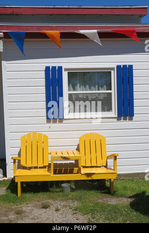 Zwei Sitzbank und Tisch im Adirondack Stil, in einem hellen Gelb, gegen ein weißes Haus mit blauen Fensterläden weatherboard Stockfoto