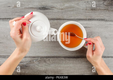 Table Top Blick auf junge Frau Hände halten weißer Keramik Teekanne in einem silbernen Löffel über heiße Tasse Tee in der anderen. Stockfoto