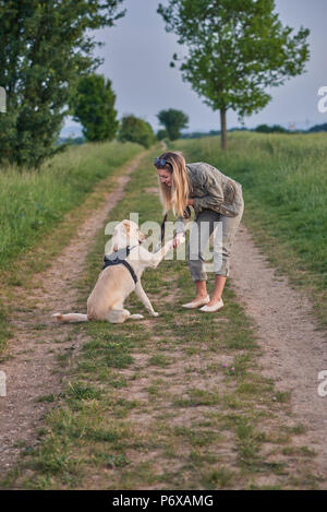 Liebevolle junge Frau eine Pfote von ihrem Hund angeboten als Sie kauert auf einem Feldweg in der Landschaft bei Ihren täglichen Spaziergang Stockfoto