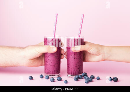 Frau und Mann hält die Hände zwei Gläser Heidelbeeren Smoothie mit Strohhalmen auf rosa Hintergrund. Gesund Sommer trinken. Stockfoto