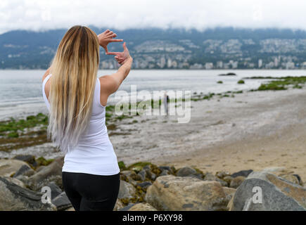 Junge Frau am Strand von Stanley Park, Vancouver im Hill Side und Framing der Ansicht mit den Händen versuchen, einen netten Platz zum Leben zu stellen. Stockfoto