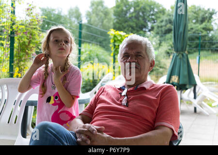 Porträt eines Mädchens und einen älteren Mann in einem Hinterhof Terrasse. Der Mann sitzt und das Mädchen stehen. Drei viertel länge. Stockfoto