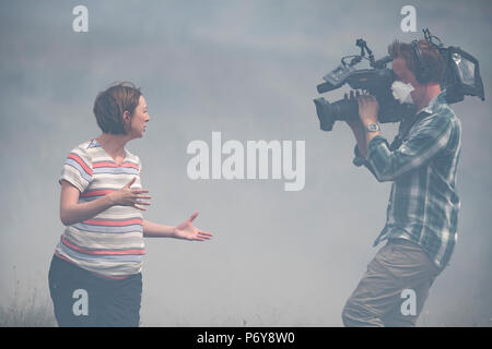 1. Juli 2018 - ein Journalist und TV-Kameramann Datensatz ein aktuelles Stück im Rauch der Winter Hill Feuer. Feuer Besatzungen aus über Großbritannien haben im Winter Hill konvergierten die Flamme, die Meter pro Stunde wachsen zu steuern. Lancashire Feuer und Rettung und Lancashire Polizei führen die Antwort auf die 'Major Incident'. Stockfoto