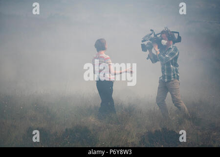 1. Juli 2018 - ein Journalist und TV-Kameramann Datensatz ein aktuelles Stück im Rauch der Winter Hill Feuer. Feuer Besatzungen aus über Großbritannien haben im Winter Hill konvergierten die Flamme, die Meter pro Stunde wachsen zu steuern. Lancashire Feuer und Rettung und Lancashire Polizei führen die Antwort auf die 'Major Incident'. Stockfoto
