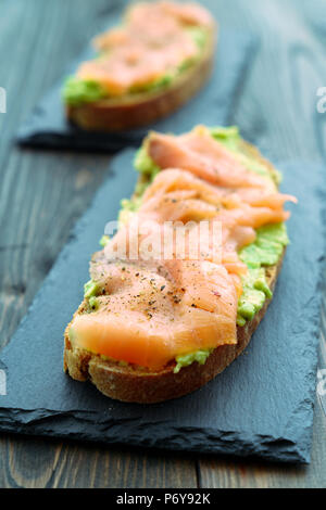 Roggen Toast mit geräuchertem Lachs und Guacamole auf schiefer Teller serviert. Hintergrund aus dunklem Holz, hohe Auflösung Stockfoto