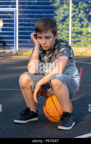 Unglückliche Junge sitzt auf einem Basketball. Spiel verlieren oder nicht für ein Team Konzept abgeholt Stockfoto