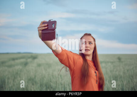 Junge schöne Frau im roten Kleid und rote Haare, nimmt eine selfie auf dem Handy in die Grüne Weizenfeld am Abend bei Sonnenuntergang im Sommer. Die Natur der digitalen Technologie. Stockfoto